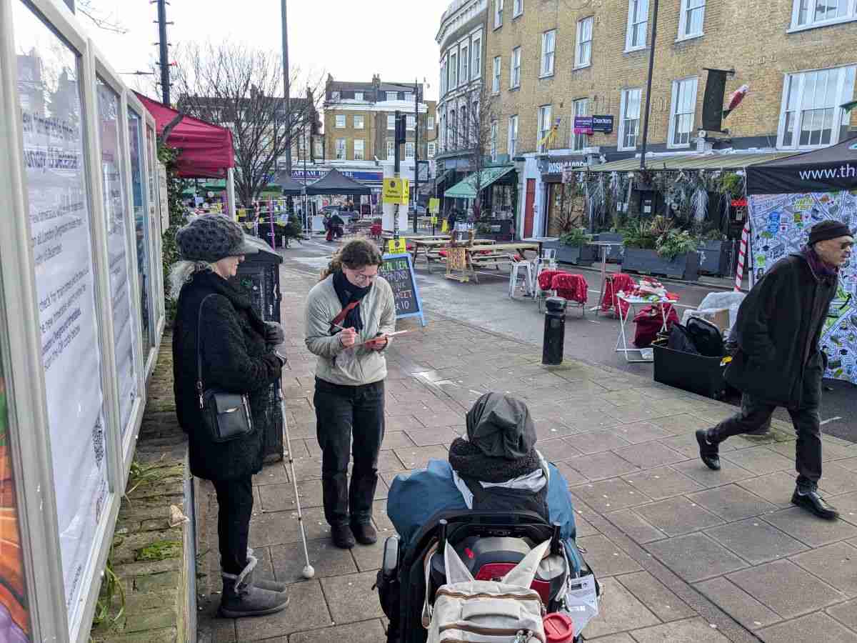 A woman standing in the street with a clipboard talking to a wheelchair user about the street environment. To the left of the photo there is a woman with a white cane. She is taking part in the conversation.