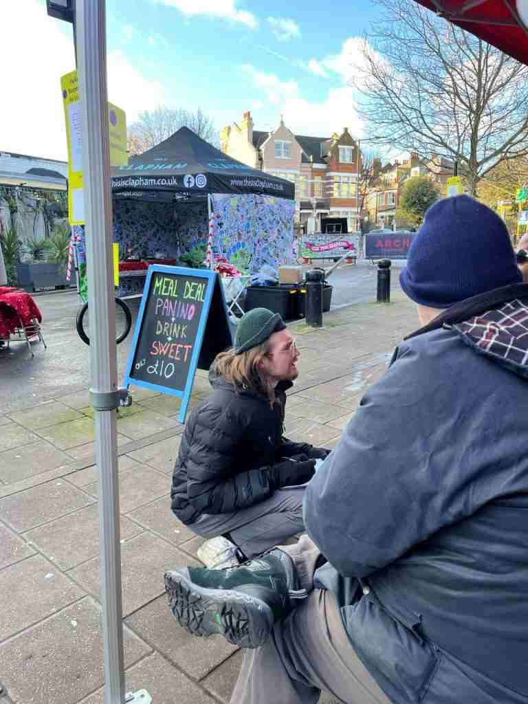 Photo shows a man talking to people who are part of the Accessibility Working Group on a street in Lambeth. There are Victorian buildings in the background and a multi-coloured gazebo and a street sign.