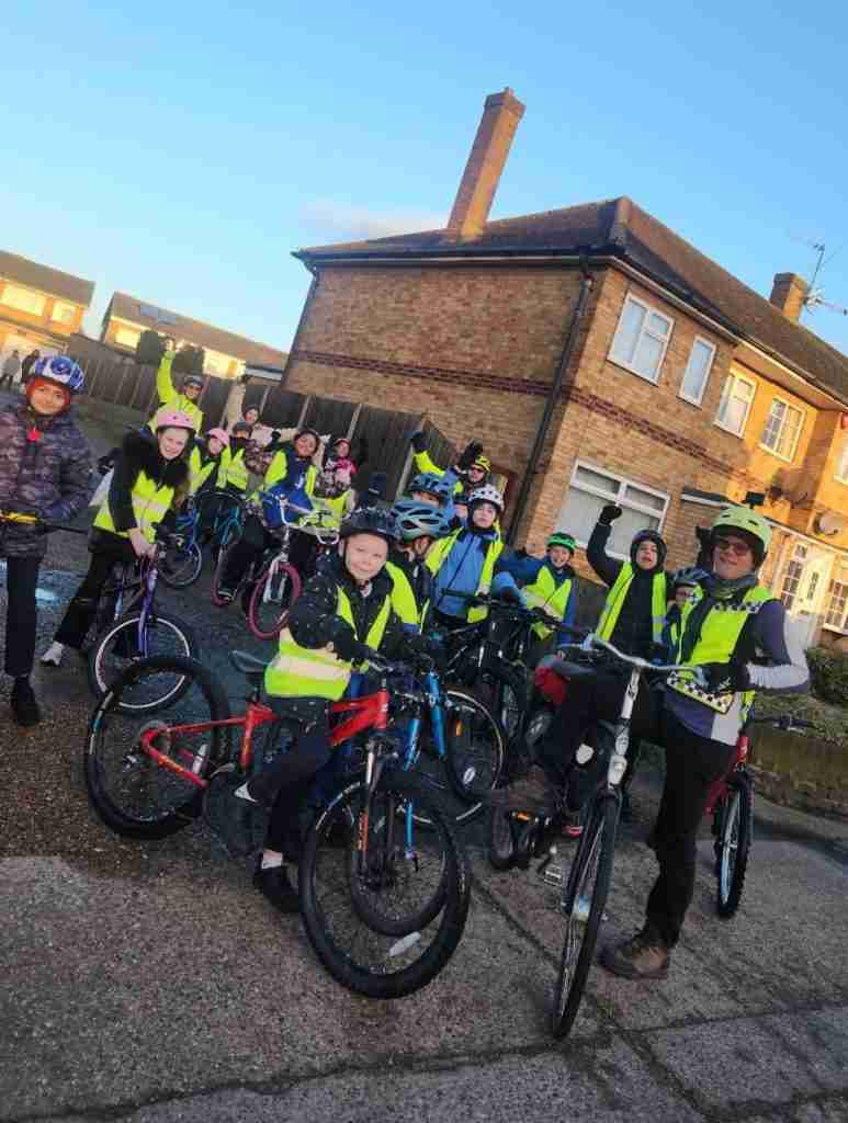 Children on the bikes looking towards the camera. They are on their way to school. They are wearing blue school uniform with hi-viz jackets.