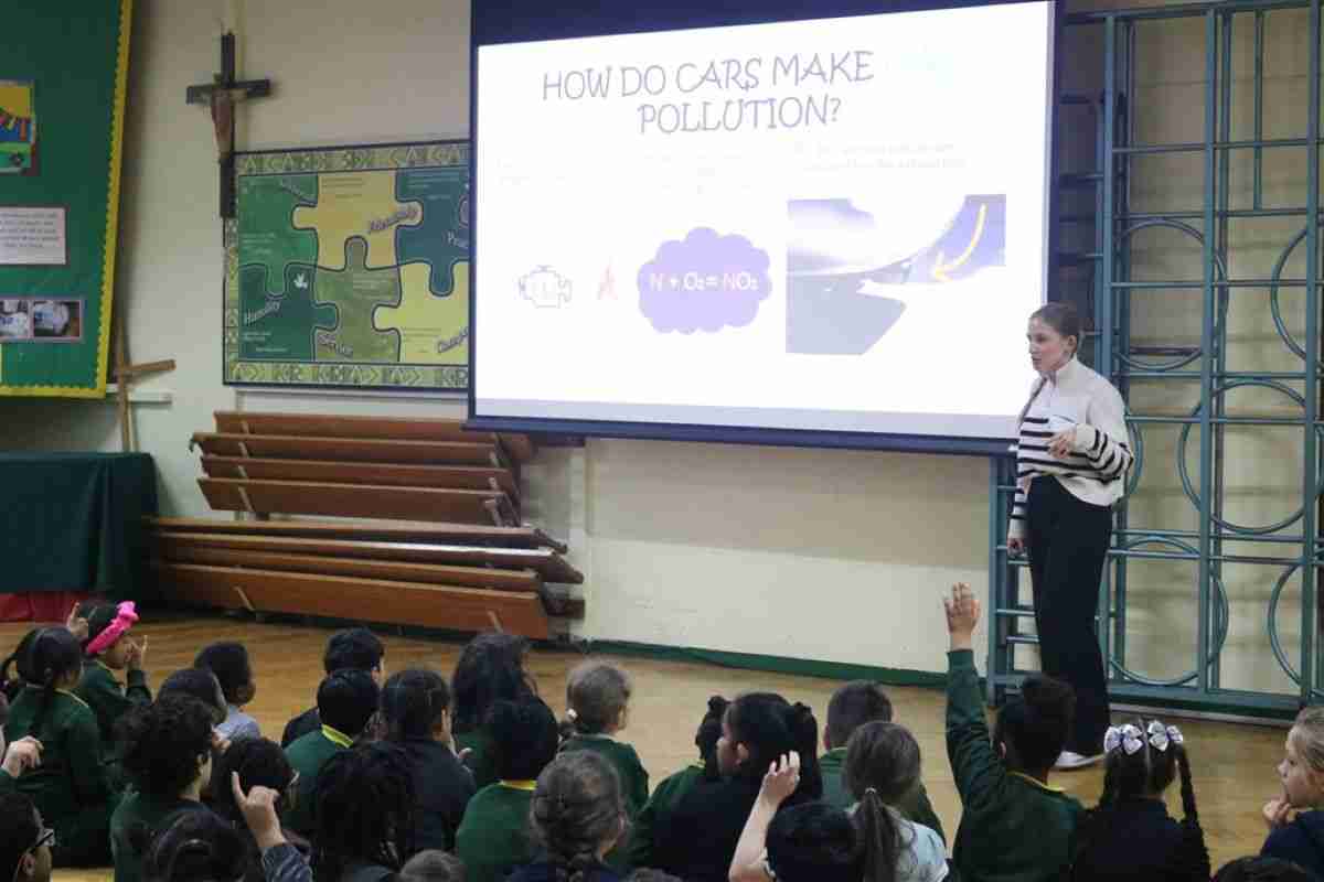 Woman standing in front of a primary school assembly. There are children sitting on the floor. Some with their hands up. the woman delivering a presentation about air pollution is talking to the children. The projector is showing a slide asking "How do cars make pollution?"