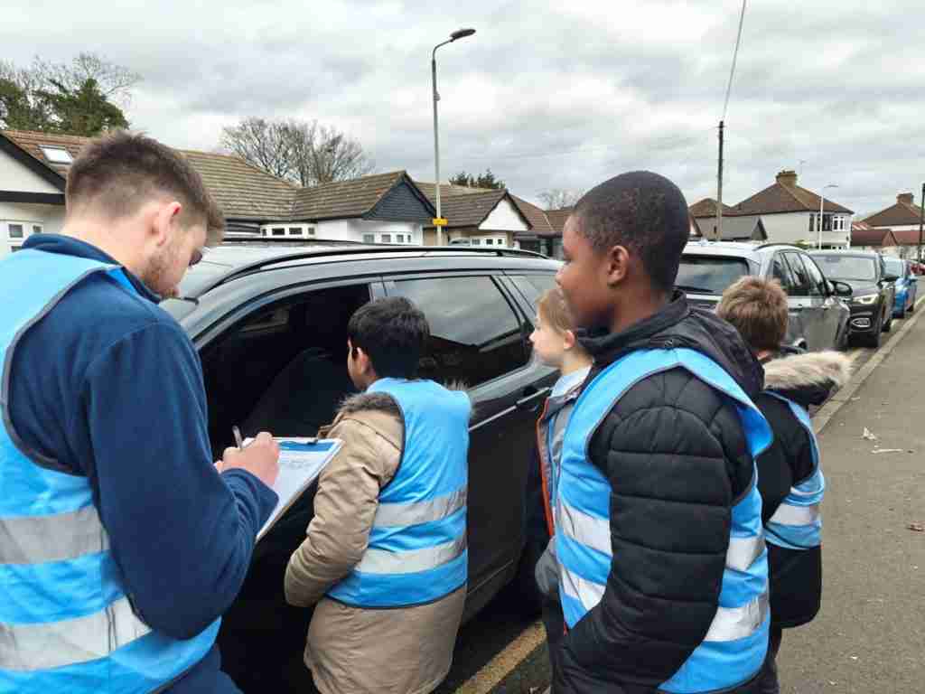 Children talking to a driver about the dangers of idling engines. The children are standing outside the car, talking to the driver through the window. They are wearing blue jackets and blue hi-vi vests.