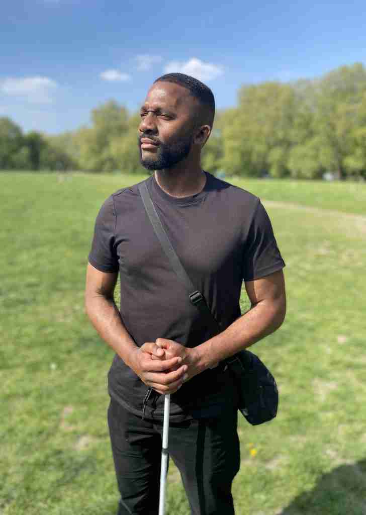 Photo shows Andre Adom of the Accessibility Working Group. He is He is wearing a dark grey t-shirt and black trousers. He has a back across him and has both hands resting on the top of his white cane. Andre is looking to the left of the viewer. In the background is blue sky with a few white clouds, green grass and trees.