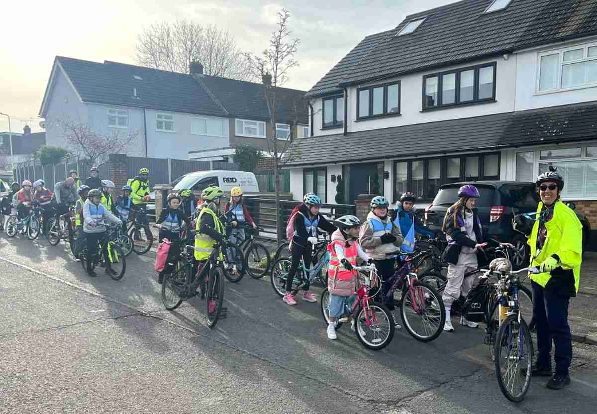 A group of children on their bikes. There is an adult cycle instructor with them. She is wearing a fluorescent jacket. The instructor and some of the children are smiling. The atmosphere is happy.