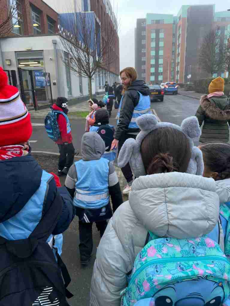 A large group of children with an adult on their way to school , They are in winter jackets and some children are carrying backpacks. The children are facing away from the camera. the adult leading them is turning around so half her face is visible.