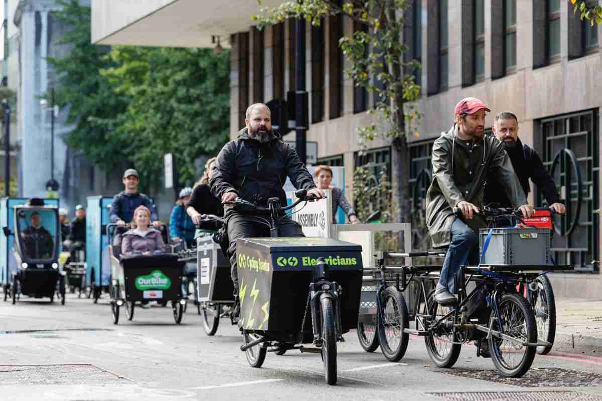 Riders and -e-cargo bikes for businesses being ridden on a London street. In the foreground is the company Cycling Sparks. The cargo bike is black with florescent yellow writing saying Cycling Sparks.