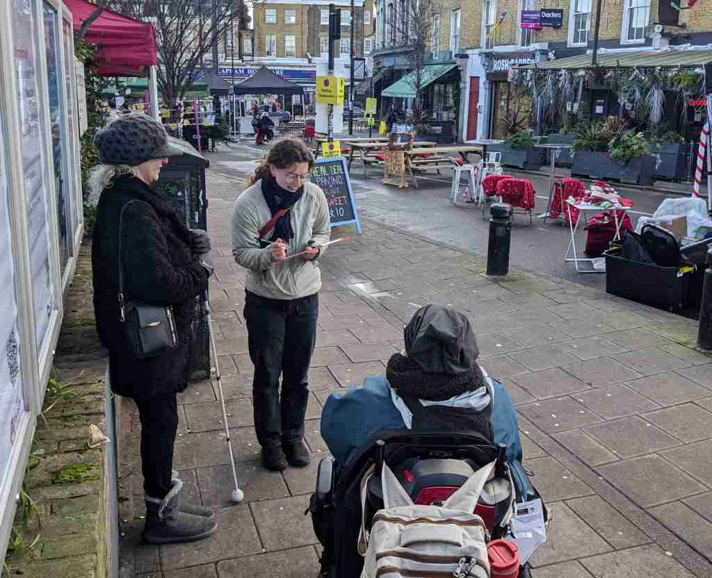 An MP Smarter Travel consultant out on the street with two members for the Accessibility Working Group. The consultant is female. She has long, curly hair and is holding a clipboard. She is us in the background. In the foreground is a woman in a wheelchair and a woman standing with a white cane. The wheelchair user has her back to the camera. The woman with the white can is standing side on.