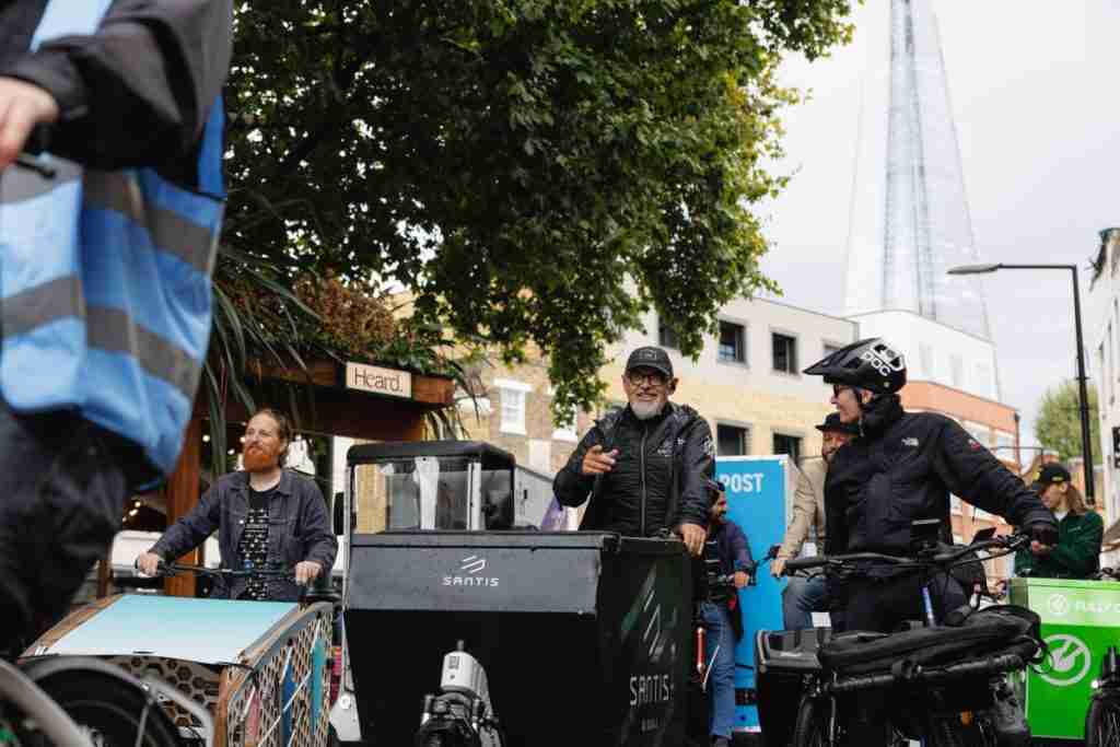 Photo shows three cargo bikes and riders. The two riders in the foreground are talking to each other and the mood of the photo is happy and positive. they are wearing black cycle clothing and riding on a city street.