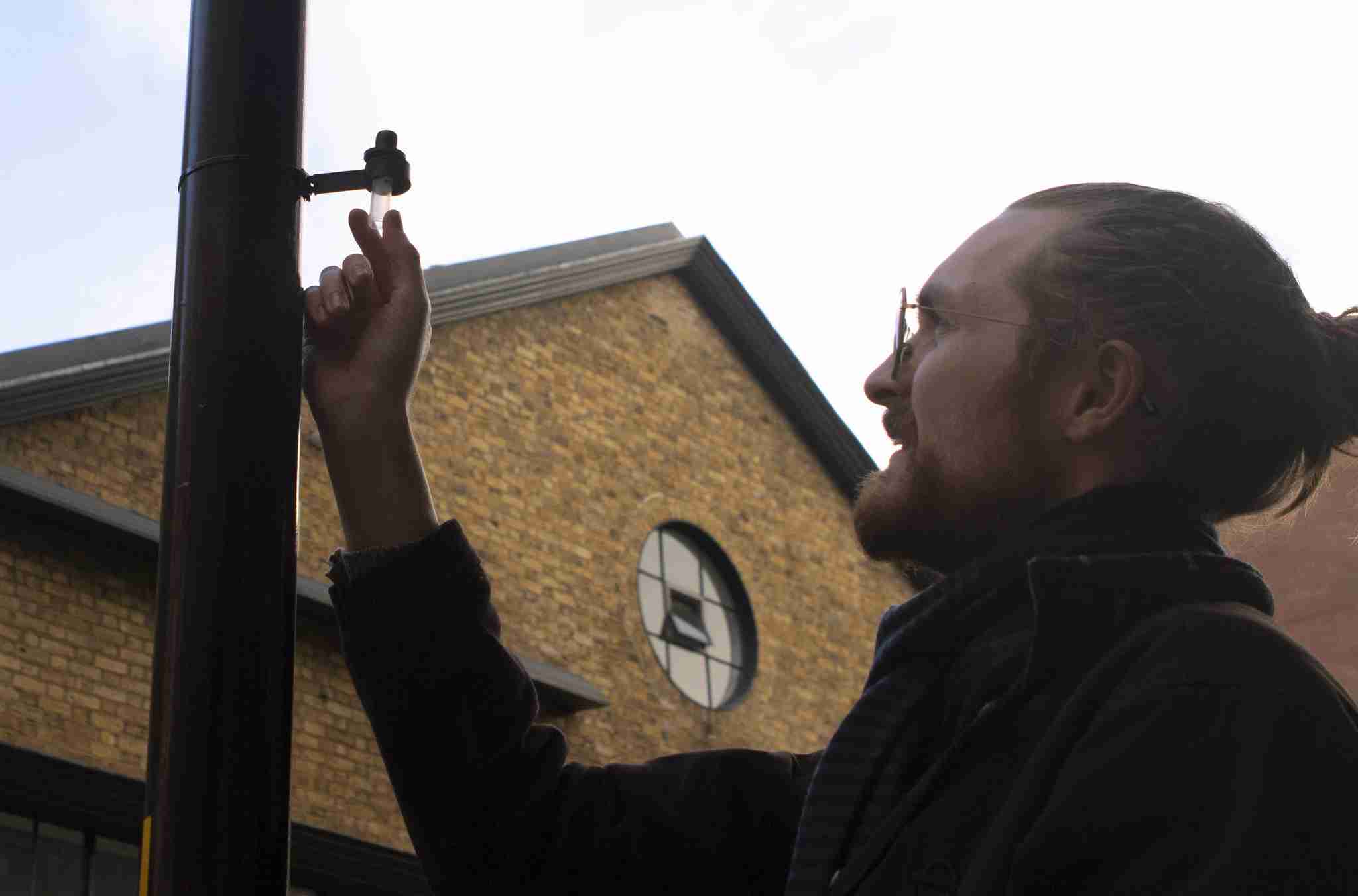 A man checking an air quality diffusion tube. The man is waring a dark brown jacket and glasses and is looking at the tube which is fixed to a lamp post.