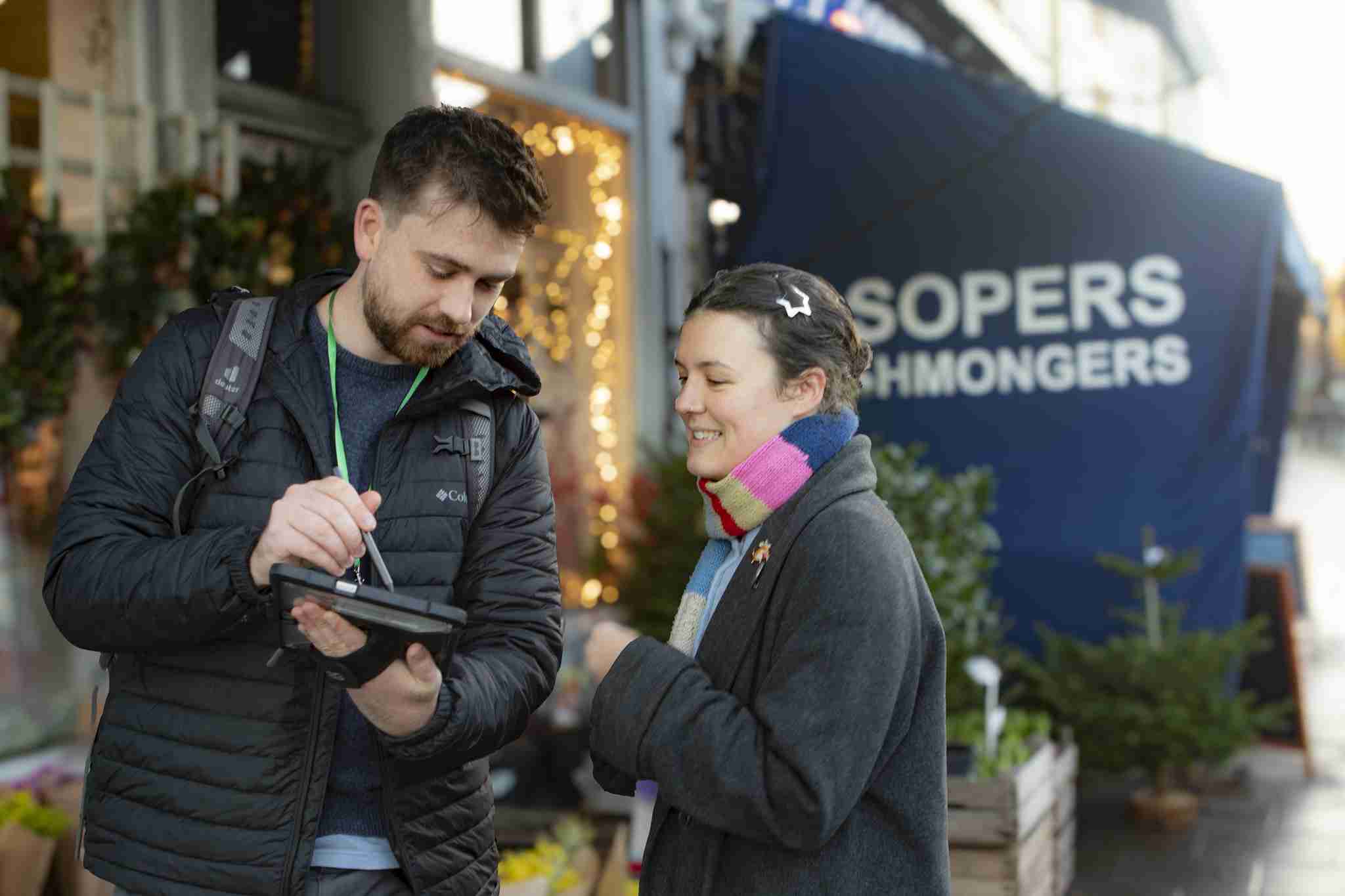 Photo shows a man and a woman in the foreground. There is a shop in the background. The man is holding a smartphone and reading out survey questions from it to the woman who is a member of the public. He is wearing a blck puffer jacket and she is wearing a dark green coat withe a bright multi-coloured striped scarf around her neck.