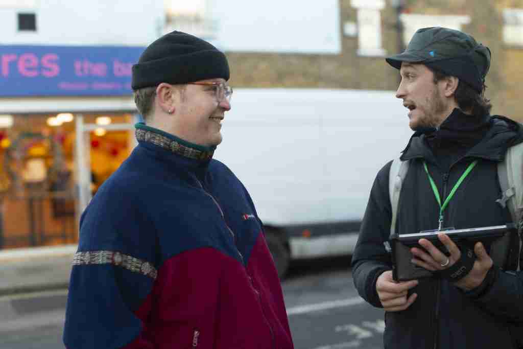 Photo shows two men in the street. The person on the right has a smart tablet and is asking the person to the left of the photo questions from his surveys.