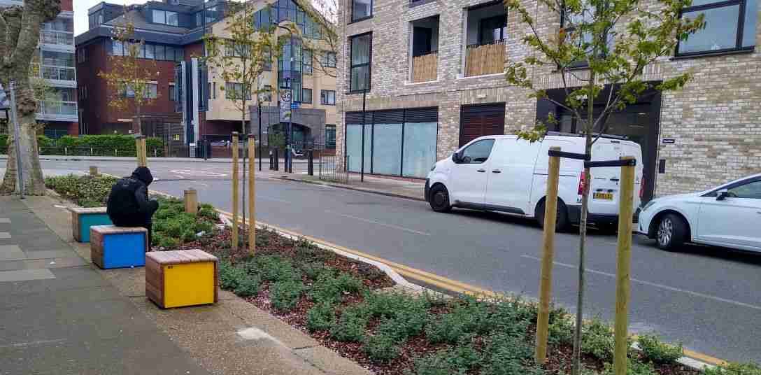 Street in London . The kerb has been built out to incorporate flower beds and trees. Seating has also been installed in the form of three yellow and blue cubes with wooden surrounds. There is a person sitting on the middle cube. They are wearing a black hoodie.