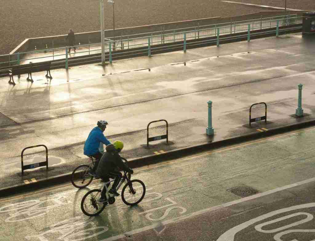Shot taken from above. Two people are cycling on a cycle lane next to each other in an urban environment. They are both wearing helmets and the road is next to the sea.