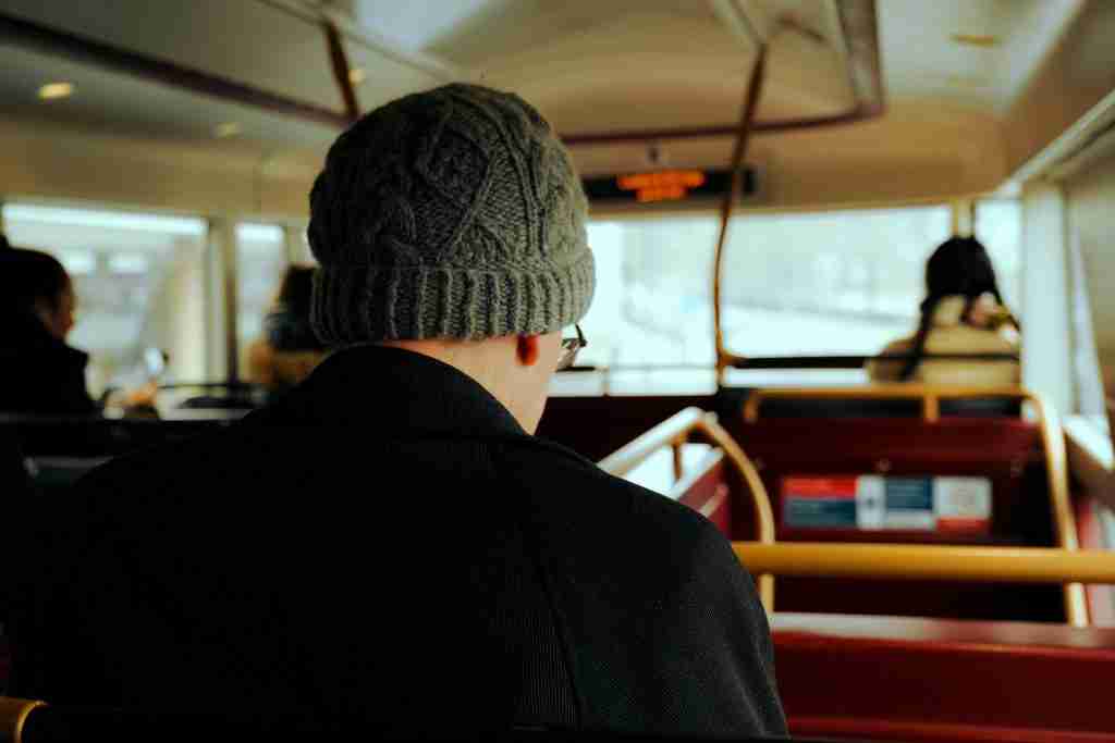 Man sitting on a bus facing away from the camera. He is wearing a green wooly hat and black jacket.
