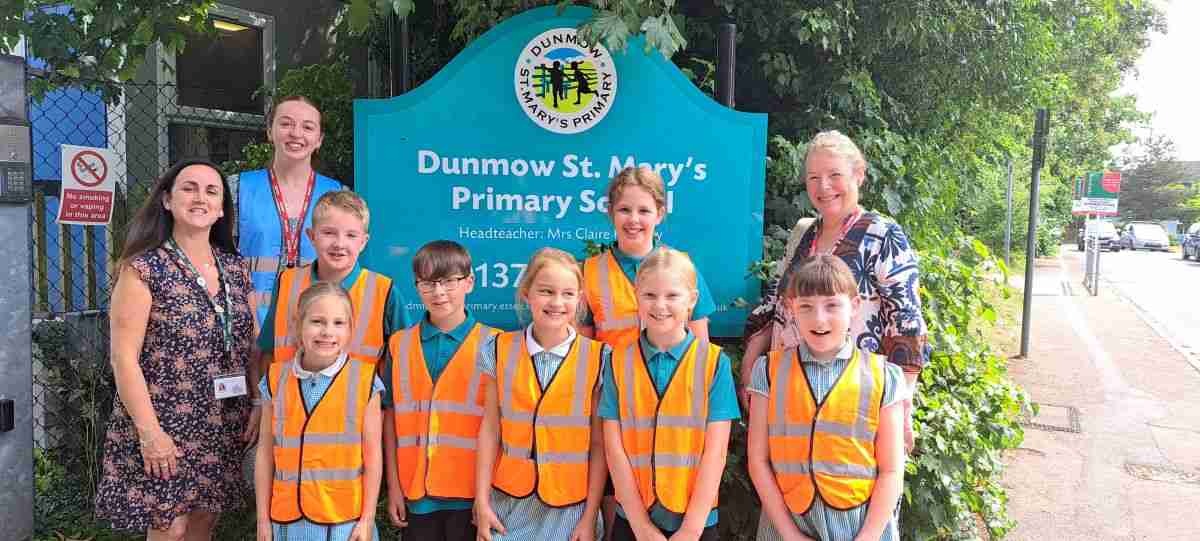 Children in school uniform wearing orange hi-z jackets. To the far left and right are teachers. They are all smiling and looking at the camera. They are taking part in the borough's Net Zero programme.