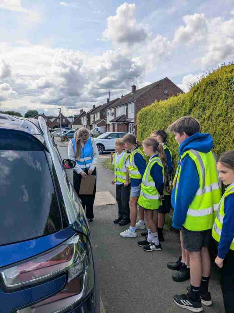 Group of six children and a teacher by a car talking to the driver about the dangers of engine idling, as part of their Net Zero programme.