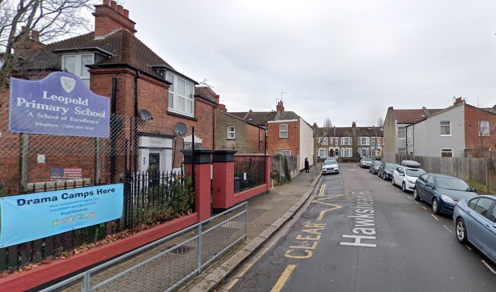 A London street with a school on it. There road has cars parked on either side. There is a person in the background wearing a black coat.