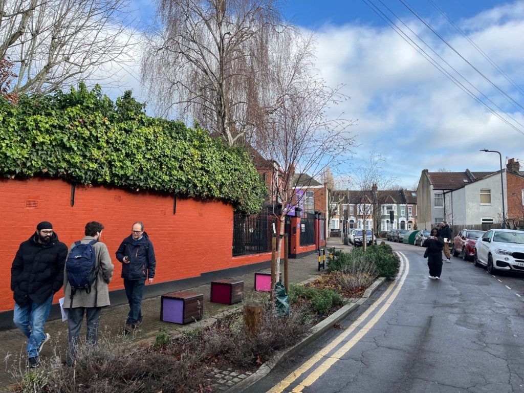 Street in Brent with kerbside planting. There are people in the street. There is also cube seating on the kerbside. They have brightly coloured sides: purple on once and pick on the others.